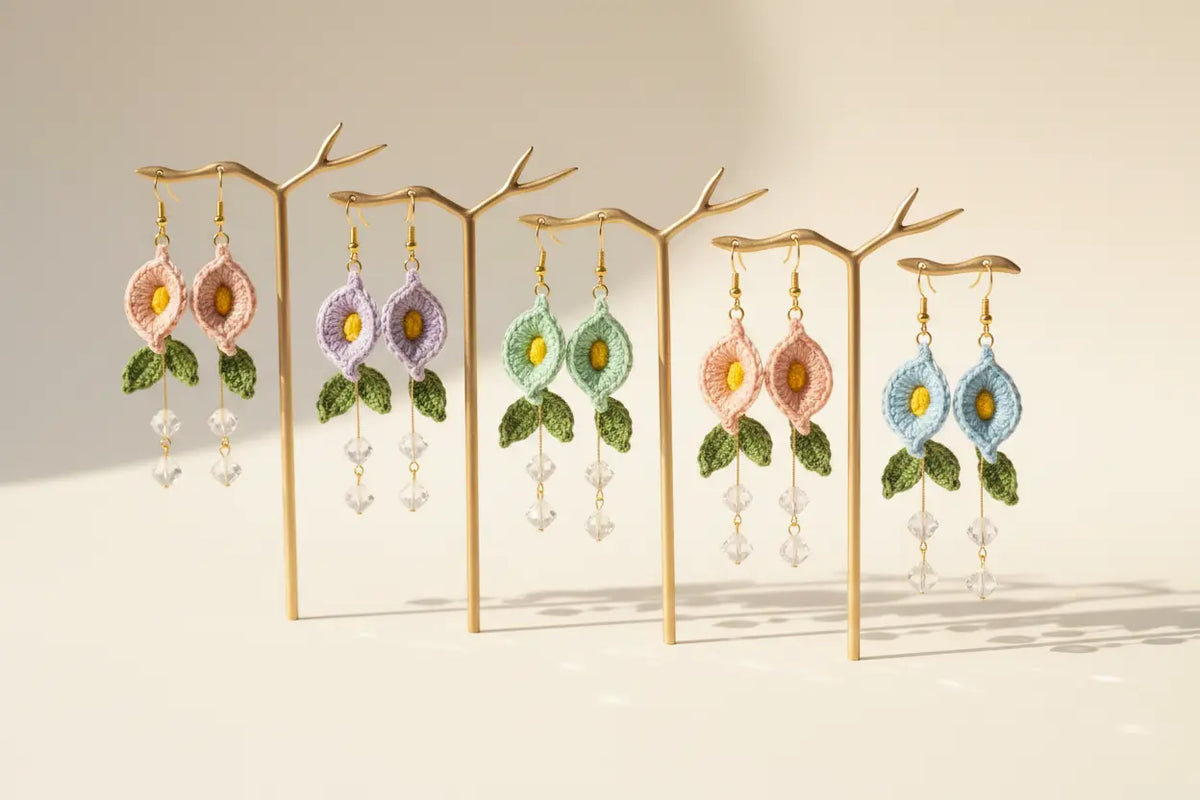 Six pairs of handmade floral dangle earrings displayed on gold branch-shaped stands against a warm beige background. The earrings feature embroidered flowers in pink, purple, mint green, peach, and light blue, with green leaves and dangling clear crystal beads.