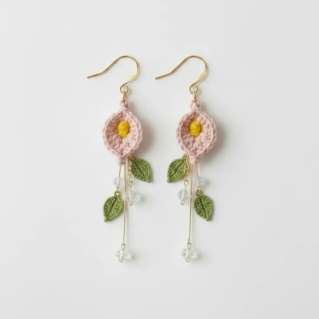 A pair of handmade crochet dangle earrings featuring light pink flower petals with yellow centers, green embroidered leaves, and gold-tone hooks and wire details, shown on a plain light gray background.