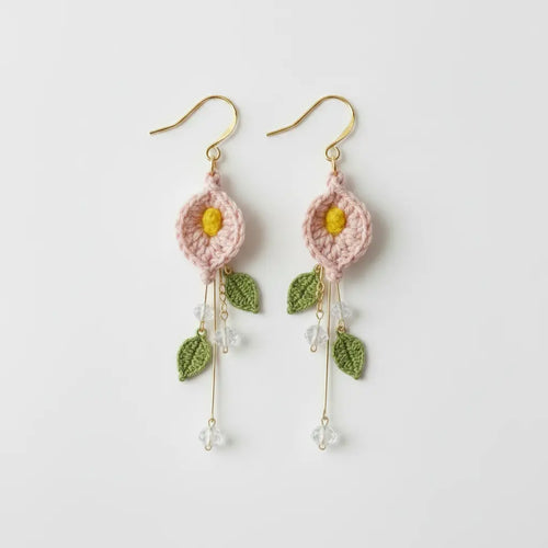 A pair of handmade crochet dangle earrings featuring light pink flower petals with yellow centers, green embroidered leaves, and gold-tone hooks and wire details, shown on a plain light gray background.