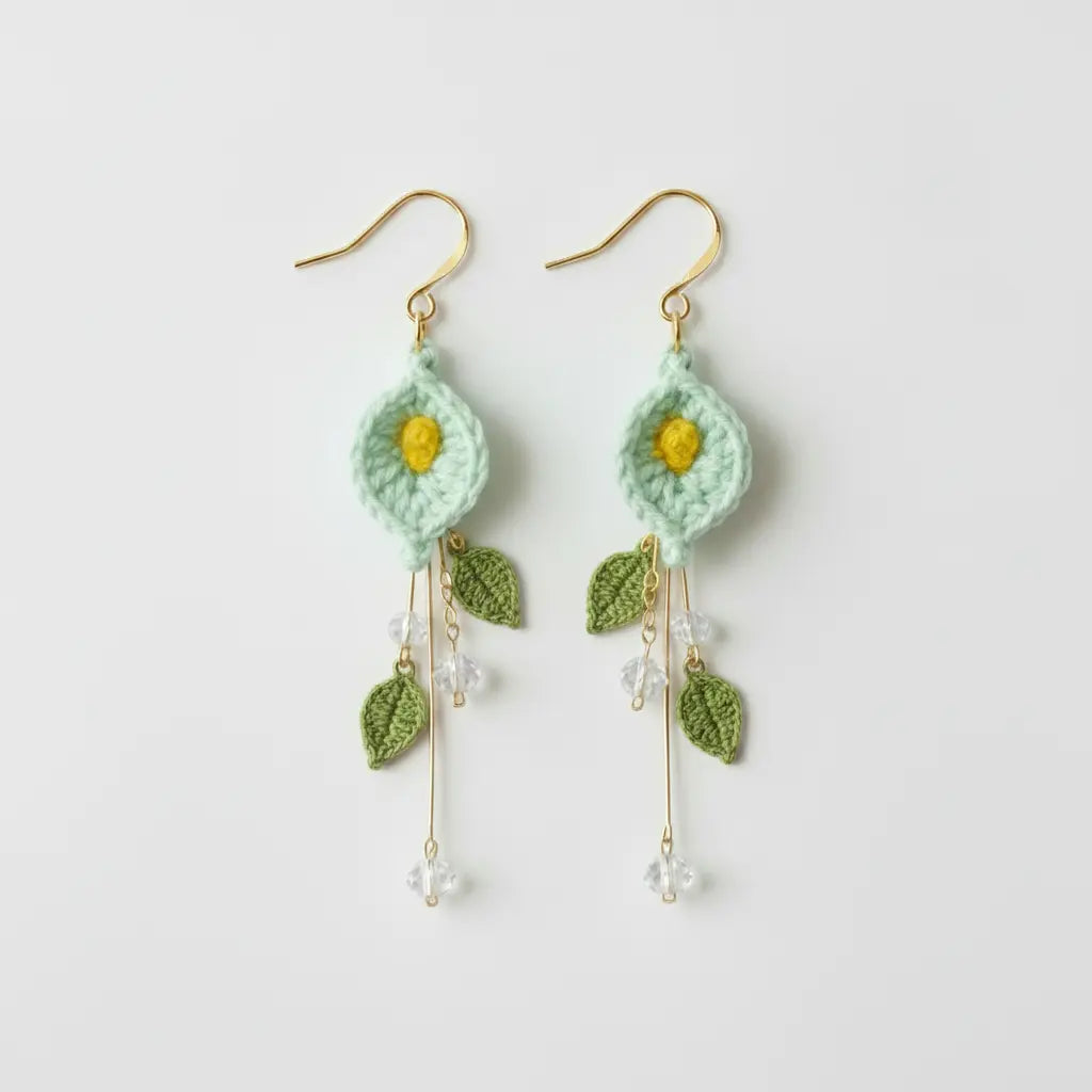 A pair of handmade crochet dangle earrings featuring mint green flower petals with yellow centers, green leaves, and gold-tone hooks and accents, shown on a plain light gray background.