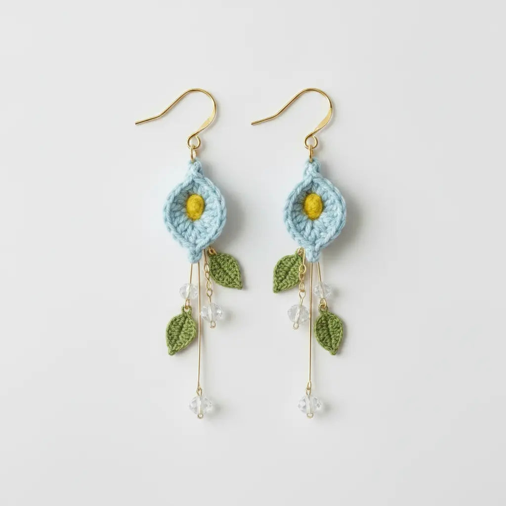 A pair of handmade crochet dangle earrings featuring light blue flower petals with yellow centers, green leaves, and gold-tone hooks on a plain light gray background.