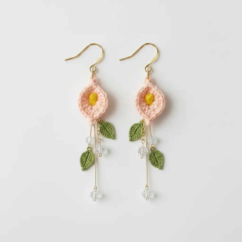 A pair of handmade crochet dangle earrings featuring light pink flower petals with yellow centers, green leaves, and gold-tone hooks and wire details, shown on a plain light gray background.