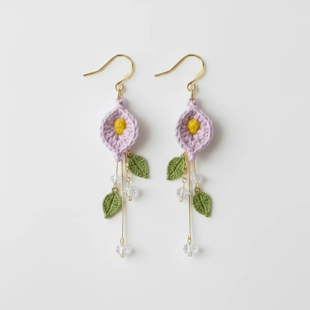 A pair of handmade crochet dangle earrings featuring light purple flower petals with yellow centers, green leaf details, and gold-tone hooks on a clean white background.