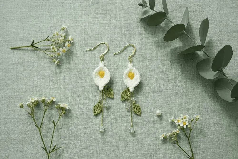 A pair of handmade crocheted daisy earrings with gold hooks, white petals, yellow centers, and green leaves, displayed on a textured sage green background with small white flowers and eucalyptus leaves.