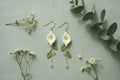 A pair of handmade crocheted daisy earrings with gold hooks, white petals, yellow centers, and green leaves, displayed on a textured sage green background with small white flowers and eucalyptus leaves.