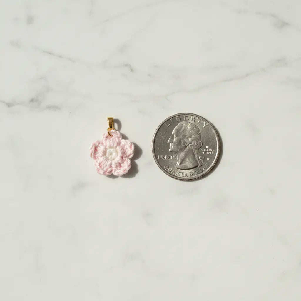 A small pink crocheted flower charm with a gold loop at the top, shown next to a US quarter coin for scale on a white marble surface.