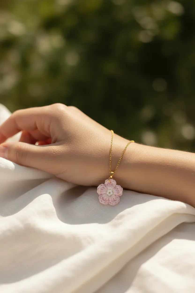 A delicate gold chain bracelet with a pink flower-shaped pendant, worn on a person's wrist resting on white fabric with a blurred green outdoor background.