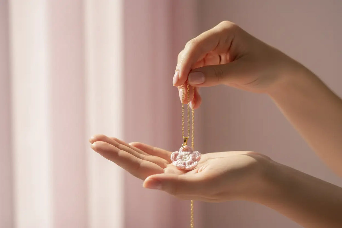 A close-up of a person's hands holding a delicate gold chain necklace with a small pink flower-shaped pendant over their open palm, set against a soft, light pink background.
