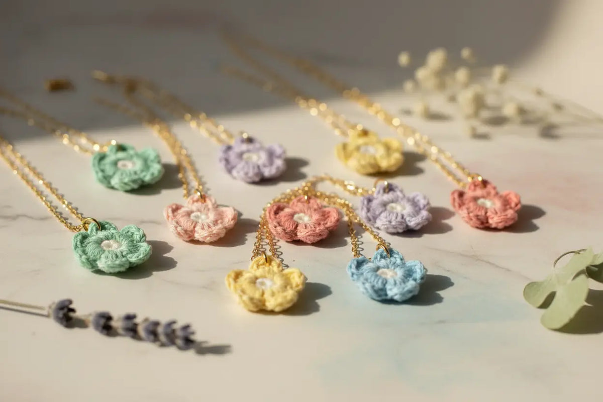 A set of pastel crocheted flower necklaces with gold chains, displayed on a light marble surface with decorative dried flowers and a green leaf.