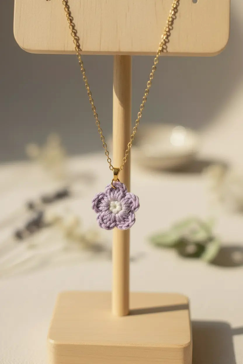 A gold chain necklace with a crocheted lavender and white flower pendant, displayed on a light wood stand against a softly blurred neutral background.