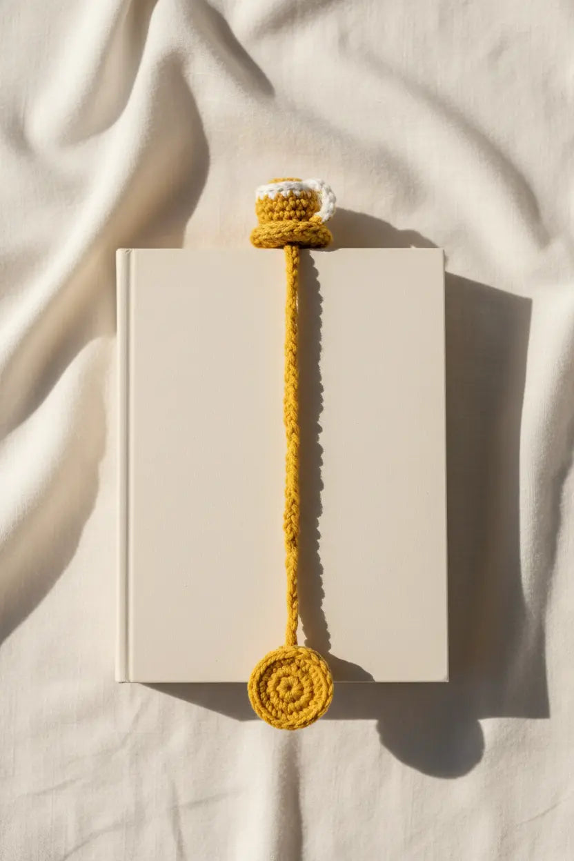 A mustard yellow crocheted book bookmark with a top loop and bottom spiral tie, shown draped over the edge of a plain white book on a cream fabric background with soft shadows.