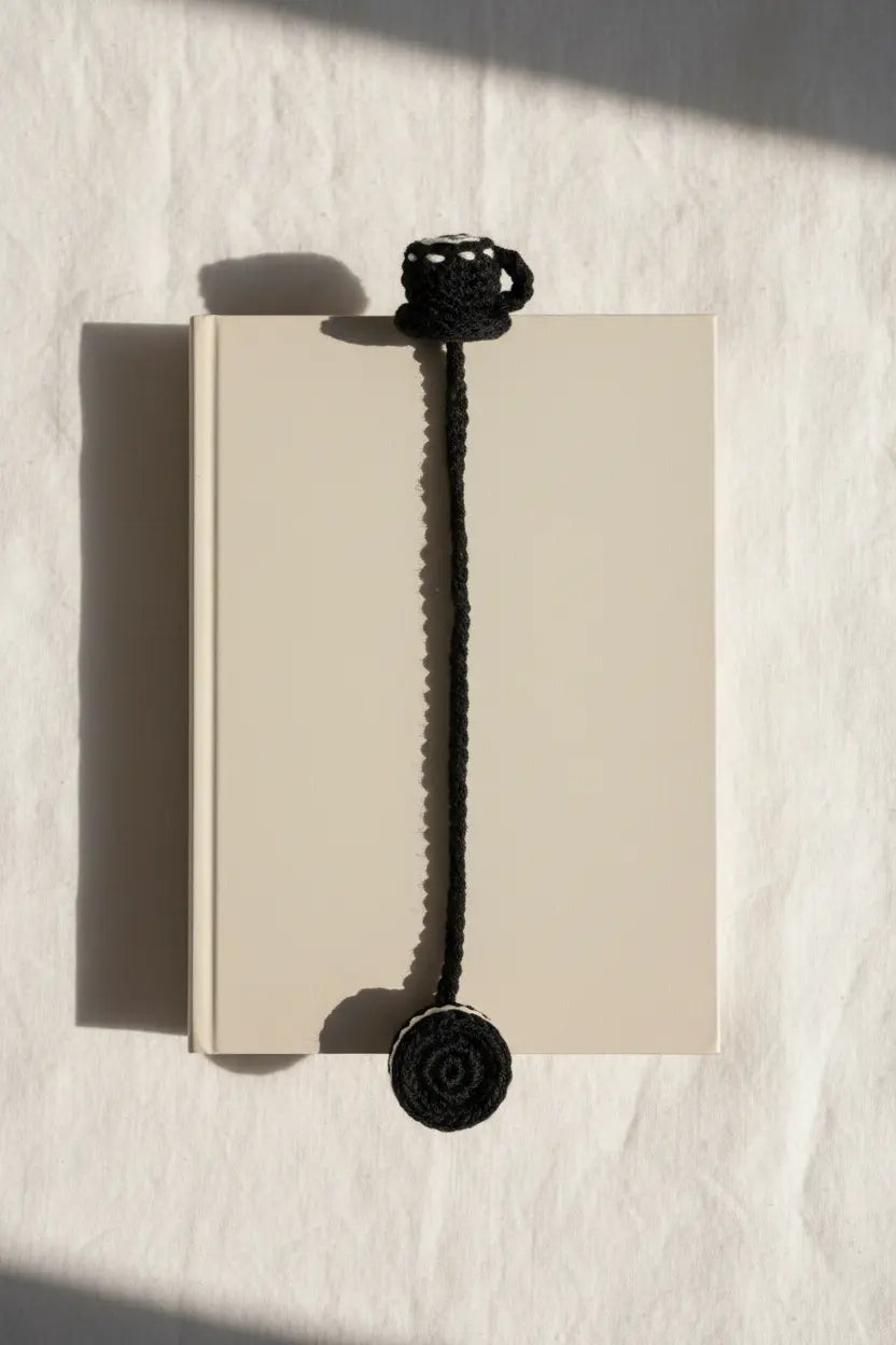 A black crocheted bookmark with a top loop and a round disc at the bottom, shown draped over a beige notebook on a white textured surface with strong sunlight and shadows.