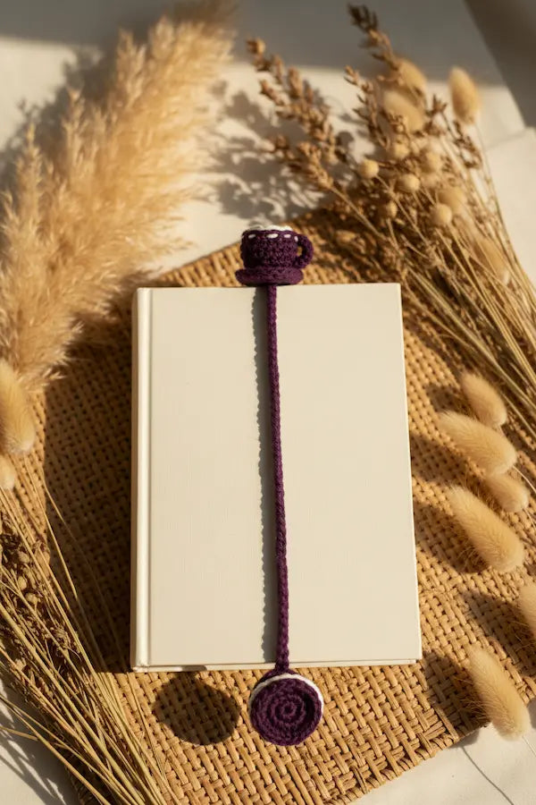 A purple crocheted book bookmark with a top loop and round spiral end, shown holding a white notebook on a woven mat with dried pampas grass in warm sunlight.