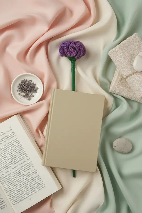 A styled flat-lay featuring a blank beige hardcover notebook with a purple knitted flower stem inserted in it, surrounded by soft draped pink and mint green fabrics, an open book, a small dish of dried lavender, and folded cream towels.