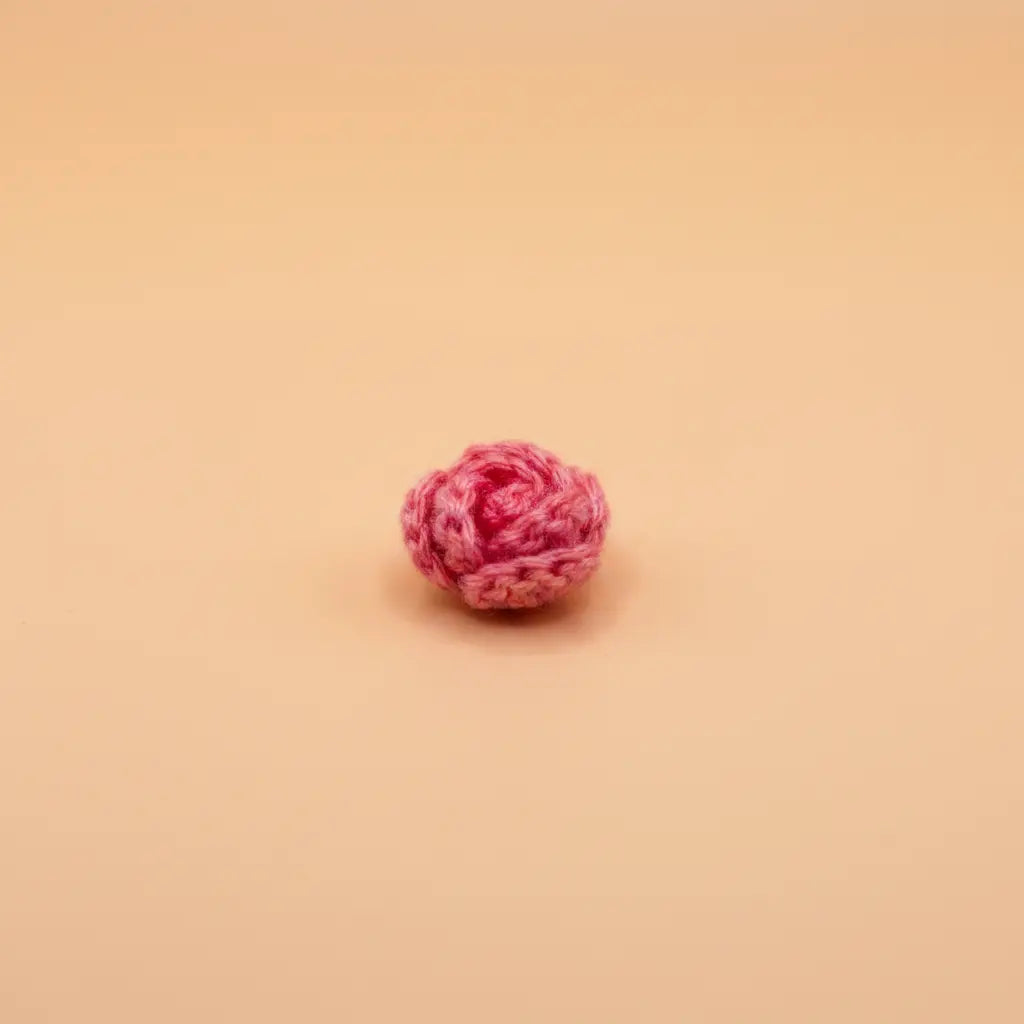 A small pink crocheted flower bud, resembling a miniature rose, centered on a solid peach-colored background.
