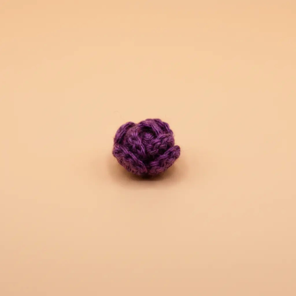 A small purple crocheted or knitted flower-shaped accessory, possibly a hair scrunchie or decorative pom-pom, centered on a solid warm beige background.