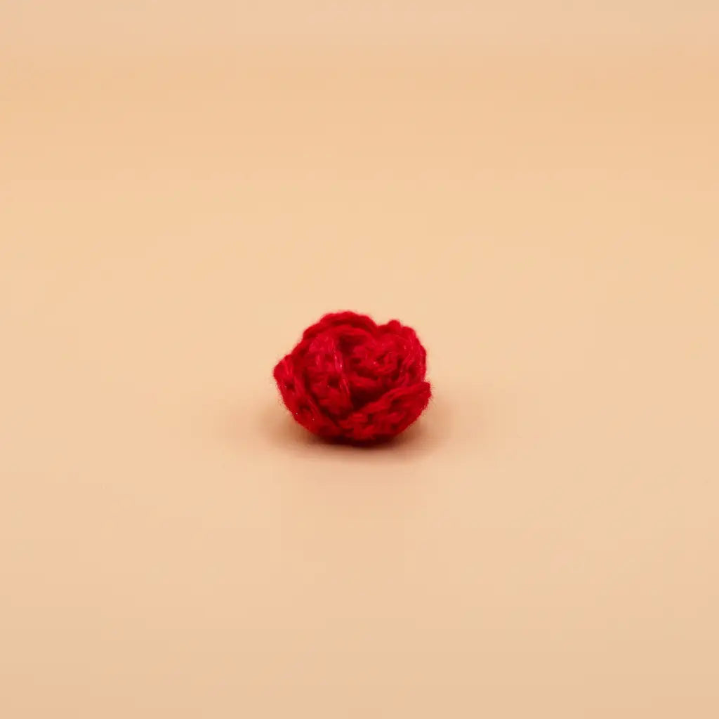 A small, vibrant red crocheted rose or flower accessory, centered on a solid, warm beige background.