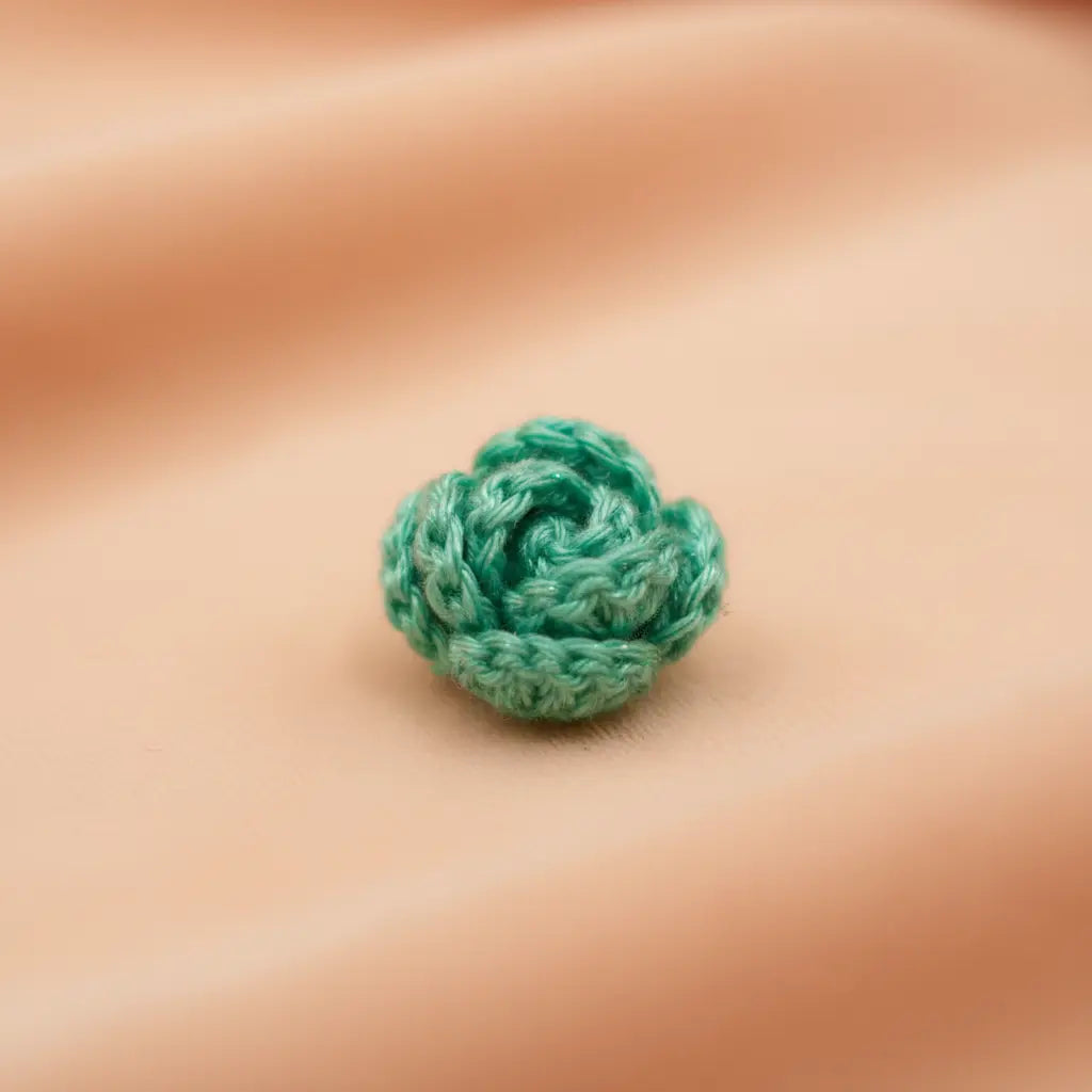 A small mint green crocheted rose-shaped embellishment or charm resting on a soft, peach-colored fabric background.