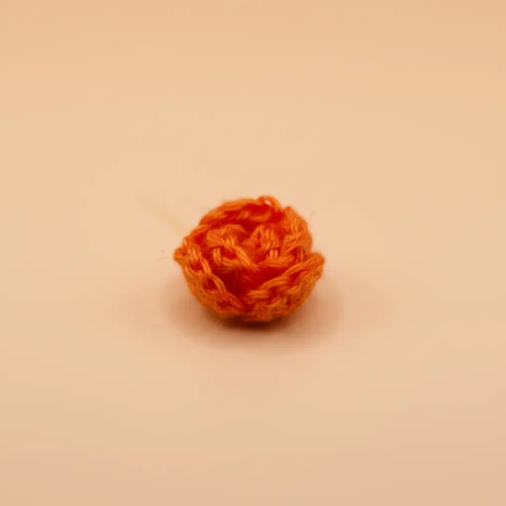 A small, textured orange crochet or knitted ball accessory, possibly a miniature pom-pom or decorative bead, shown on a plain beige background.