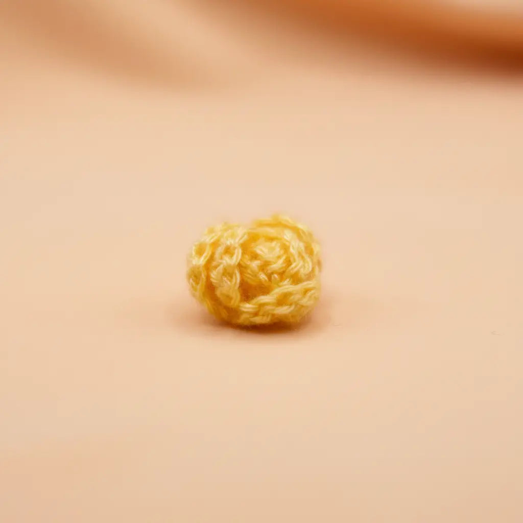 A small, round, yellow crocheted or knitted textured bead or charm, centered on a smooth, warm beige background.