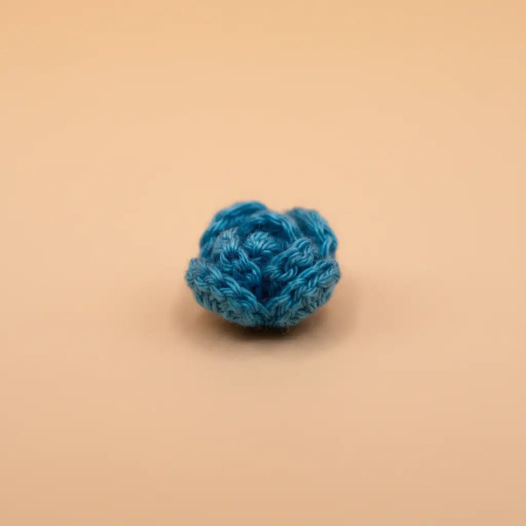 A small crocheted blue flower-like pom-pom or decorative ball made from textured yarn, centered on a smooth warm beige background.