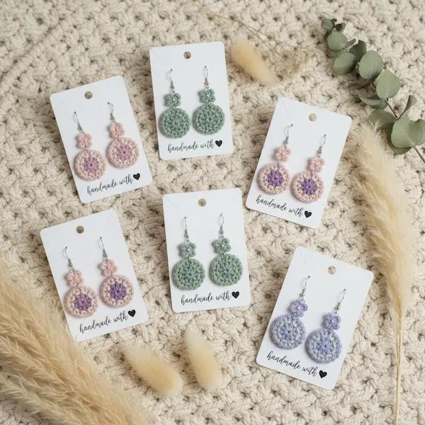 Five pairs of handmade crocheted dangle earrings displayed on white cards over a beige knit blanket, with dried grass and eucalyptus leaves around them. The earrings feature round and oval crochet motifs in pink, green, and lavender.