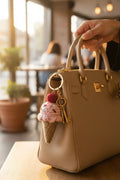 A beige pebbled leather handbag with gold hardware, held by a hand in a warm, softly blurred indoor setting. A crocheted pink and beige ice cream cone keychain charm with a gold key hangs from the bag's ring.
