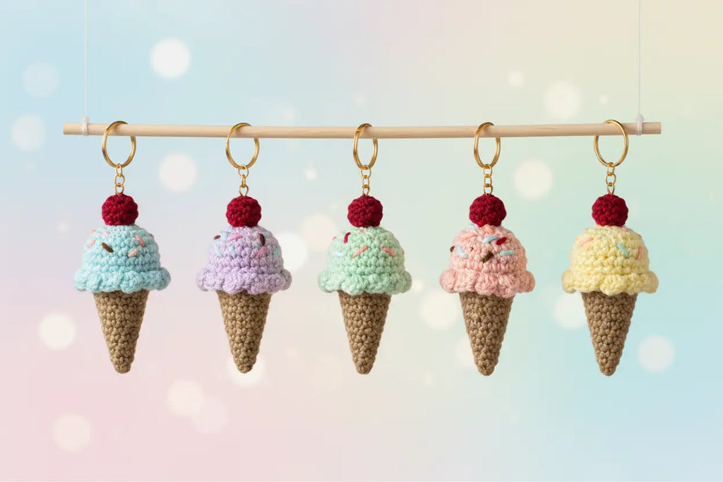 Five crocheted ice cream cone keychains in pastel shades of blue, purple, mint green, pink, and yellow, hanging from a wooden rod against a soft pastel bokeh background.