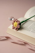 A crocheted flower bookmark in dusty pink and yellow rests on an open book, with a green stem and ribbon on a soft pink background.