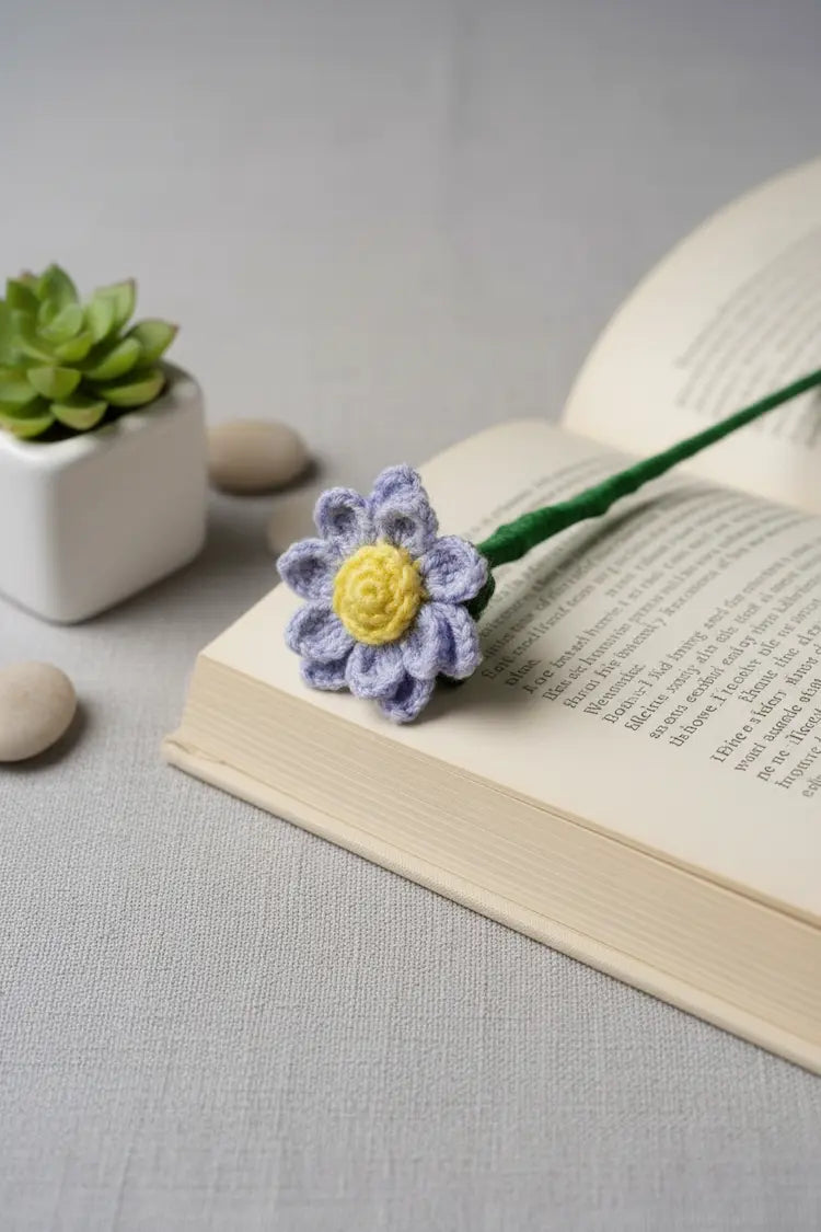 A handmade crochet flower bookmark with a lavender-purple and white petals and a yellow center, resting on an open book. The book stem is green, and the scene is styled with a small potted succulent and pebbles on a textured gray surface.
