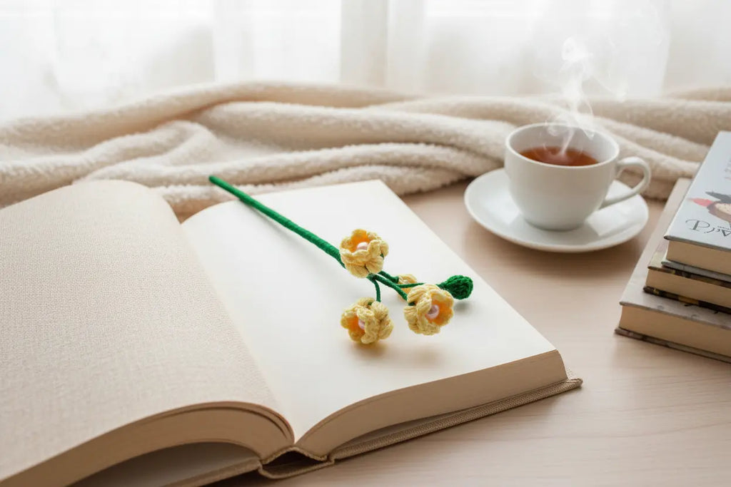 A handmade crocheted book mark shaped like yellow flowers on a green stem rests on an open book on a wooden table. In the background, there is a cup of tea on a saucer, a stack of books, and a soft beige blanket.