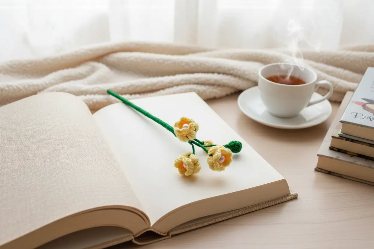 A handmade crocheted book mark shaped like yellow flowers on a green stem rests on an open book on a wooden table. In the background, there is a cup of tea on a saucer, a stack of books, and a soft beige blanket.