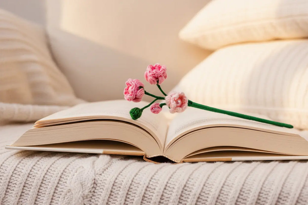 An open book on a cream knit blanket with soft pillows in the background, and a small pink flower with a green stem resting across the pages.