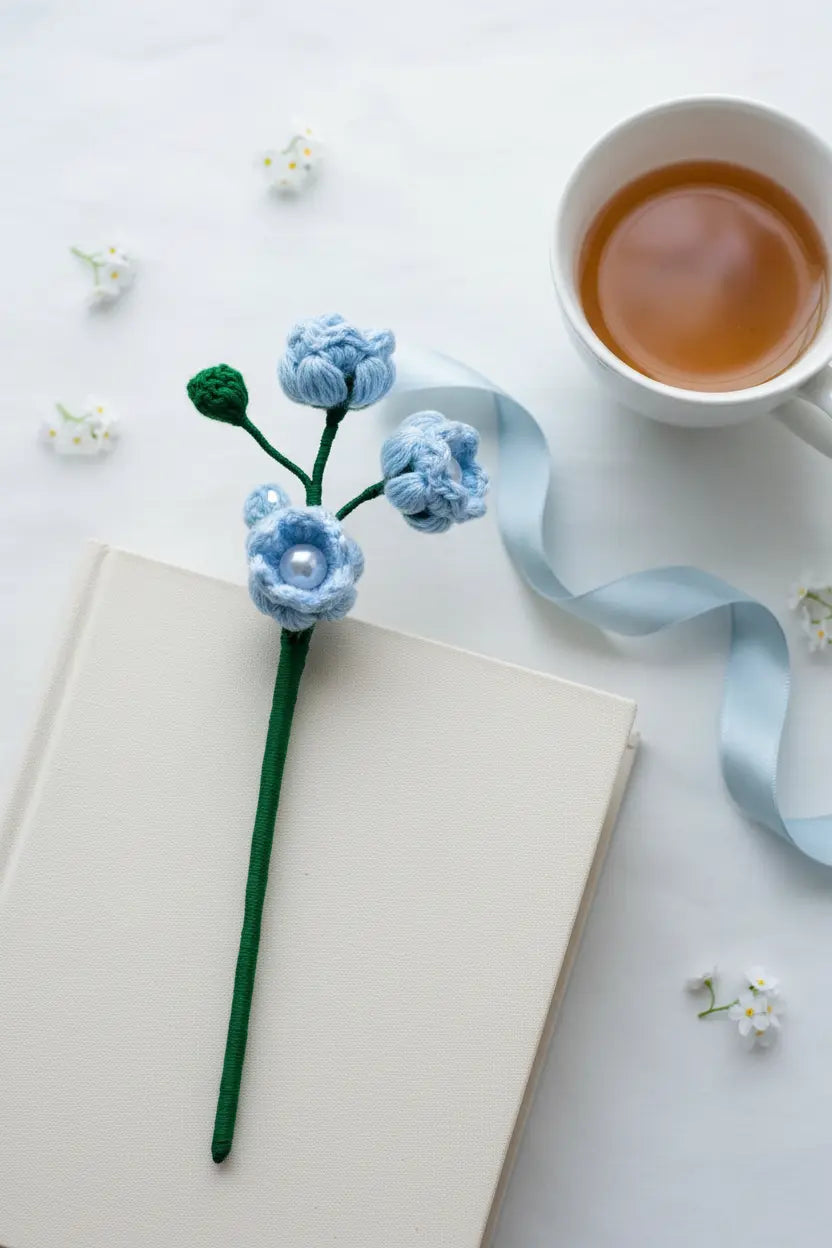 A handmade crochet flower bookmark with light blue blossoms and a green stem rests on an open cream-colored book. The scene is styled with a white cup of tea, a light blue ribbon, and small white flowers on a white marble surface.
