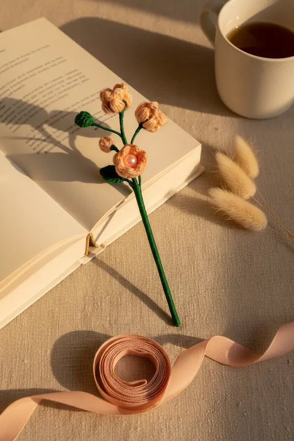 A handmade crocheted flower bookmark with peach-colored blossoms and a green stem rests on an open book. The scene is styled with a roll of peach ribbon, dried bunny tail grass, and a white mug of tea on a beige fabric surface.