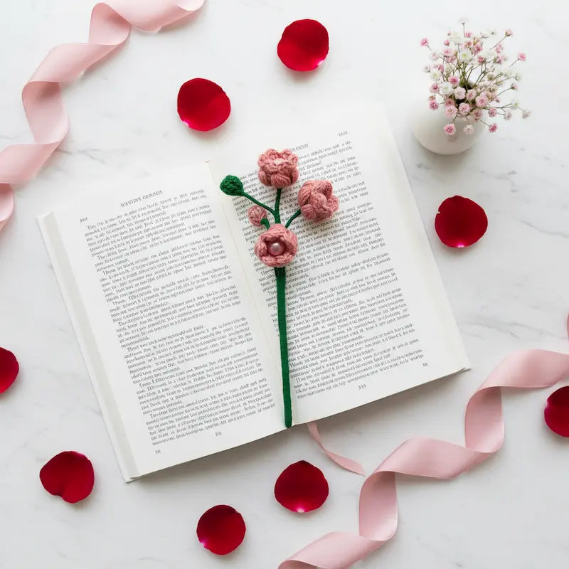 A pink handmade crochet flower bookmark with a green stem rests on an open book, surrounded by pink ribbons, red rose petals, and a small vase of baby's breath on a white marble surface.