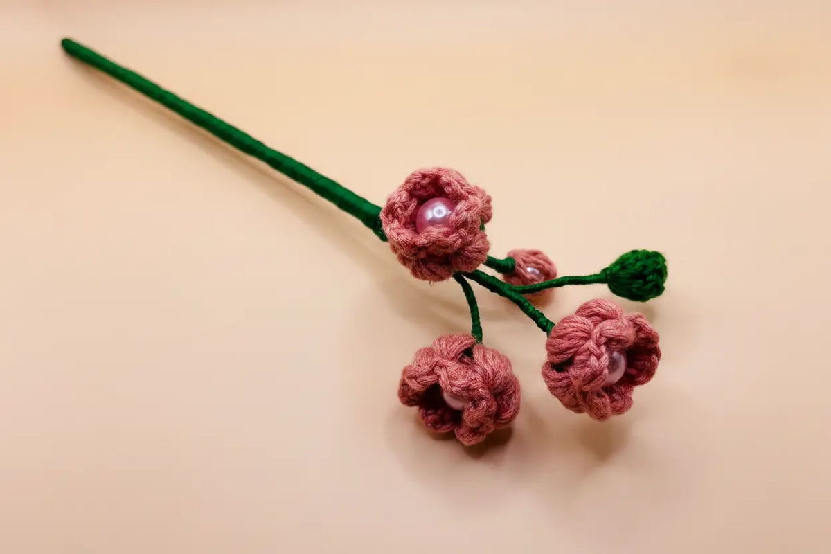 A handmade crochet hair stick featuring three pink crocheted flower buds with pearl-like centers, green yarn stems, and a small green leaf accent, displayed on a warm beige background.