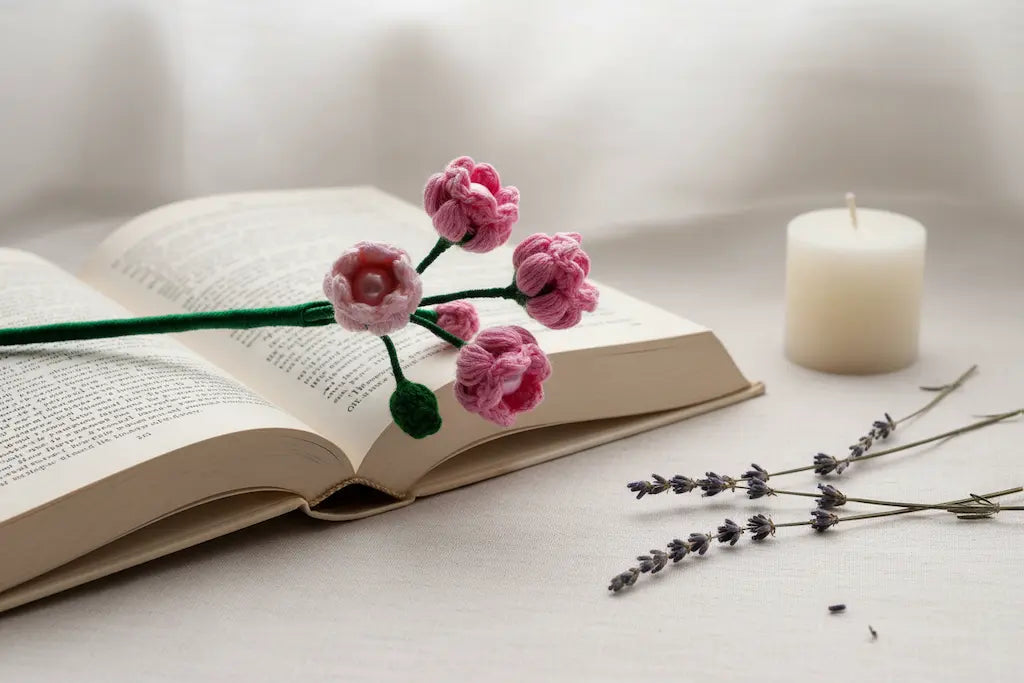A pink and light pink crochet flower stem with green stems and leaves rests across an open book, styled on a light surface with a white candle and dried lavender sprigs nearby.