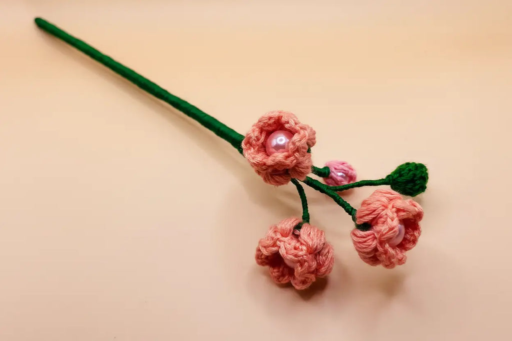 A handmade pink crocheted floral hair accessory with a green stem, three pink flower buds, and small pearl-like center accents, displayed on a light beige background.