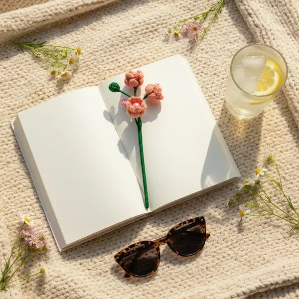 A styled flat-lay featuring a white open book with a pink flower-shaped bookmark on the page, surrounded by small daisy flowers, tortoiseshell sunglasses, and a glass of lemonade on a beige knit blanket.