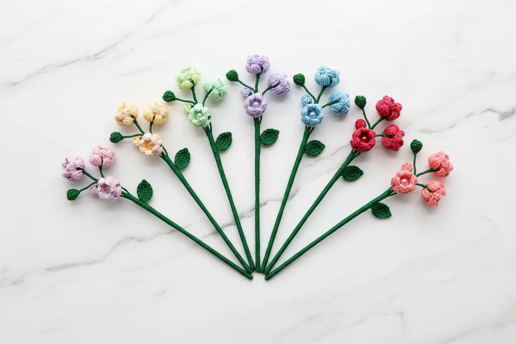 Seven mini crochet flowers with green stems and leaves, arranged in a fan shape on a white marble background. The flowers come in pink, peach, yellow, mint green, lavender, blue, and red.