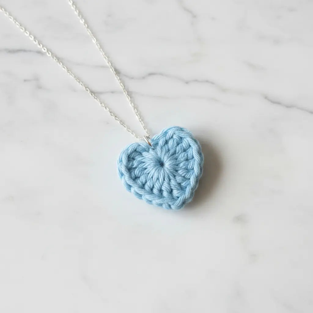 A light blue crocheted heart pendant necklace on a delicate silver chain, displayed on a white marble surface.