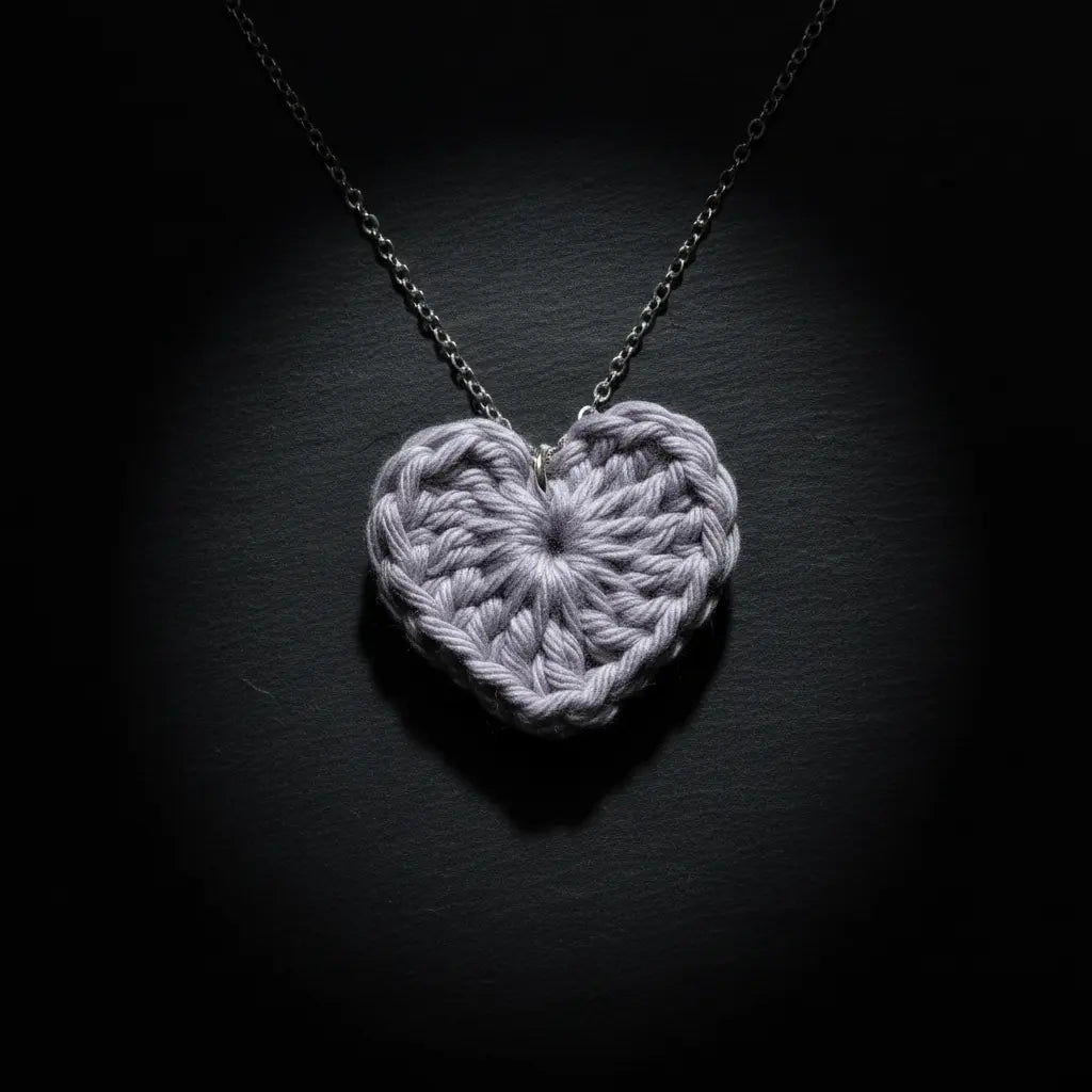 A light gray crocheted heart pendant necklace on a silver chain, displayed against a dark, spotlight-lit background.