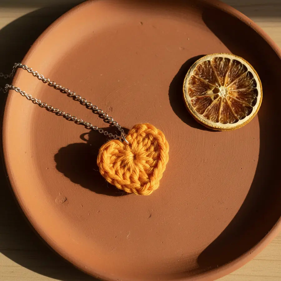 Crocheted orange heart pendant on a chain next to a dried orange slice on a brown plate.
