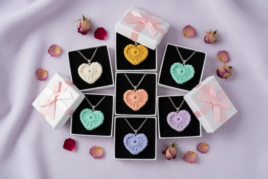 Six heart-shaped crochet pendant necklaces in pastel colors, each displayed in an open black jewelry box with a white gift box and pink ribbon on a light lavender fabric background, surrounded by scattered rose petals.