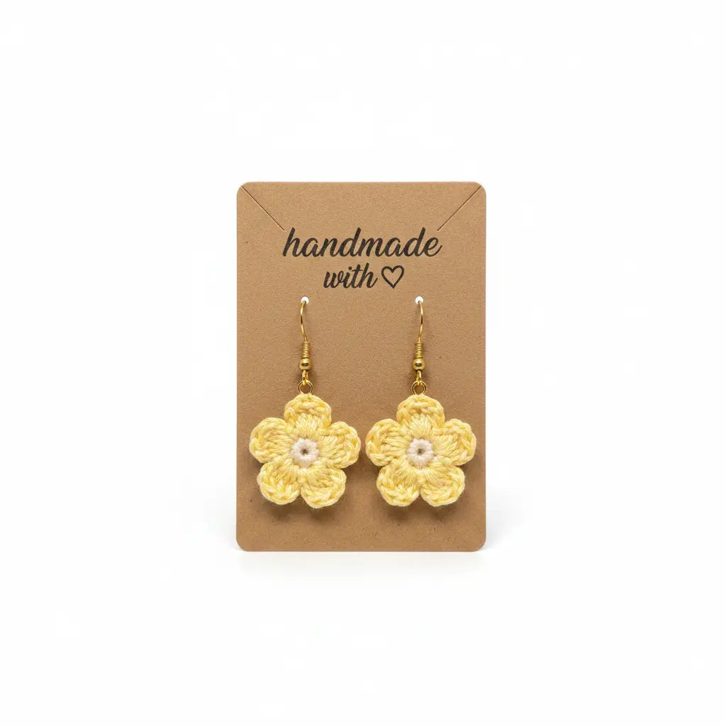 A pair of handmade yellow crocheted flower dangle earrings with white pearl-like centers and gold hooks, displayed on a brown kraft card that reads 'handmade with heart' against a white background.