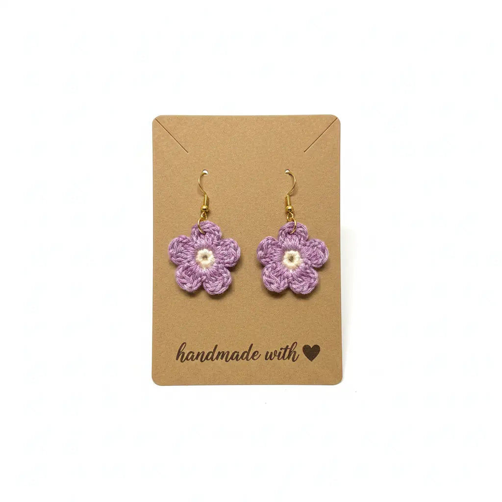 A pair of handmade crochet flower earrings in light purple with a small white center and gold hooks, displayed on a brown kraft card that reads 'handmade with heart'.