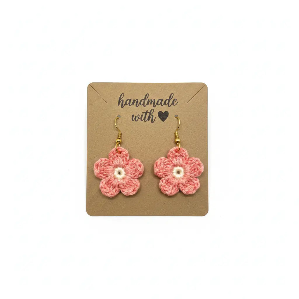 A pair of handmade crocheted flower drop earrings in pink with white centers and gold hooks, displayed on a brown kraft card that reads 'handmade with a heart'.