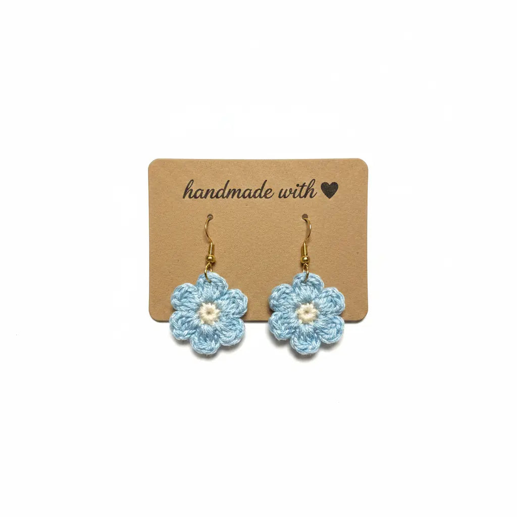 A pair of handmade light blue crocheted flower dangle earrings with white centers and gold hooks, displayed on a brown kraft card that reads 'handmade with' against a white background.