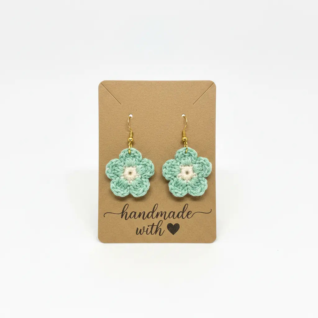 A pair of handmade crocheted flower dangle earrings in mint green and white, displayed on a brown kraft card that reads 'handmade with' against a plain white background.